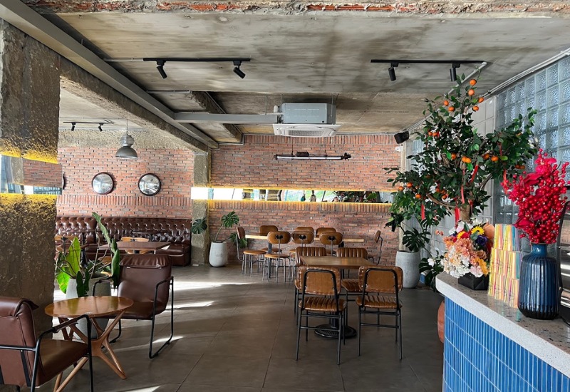 Qu&aacute;n c&agrave; ph&ecirc; với b&agrave;n ghế gỗ, tường gạch th&ocirc;, trang tr&iacute; c&acirc;y xanh v&agrave; hoa, &aacute;nh s&aacute;ng tự nhi&ecirc;n chiếu v&agrave;o.