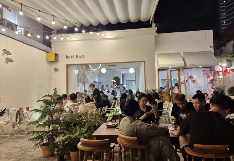 Qu&aacute;n c&agrave; ph&ecirc; ngo&agrave;i trời đ&ocirc;ng kh&aacute;ch v&agrave;o buổi tối, với &aacute;nh đ&egrave;n trang tr&iacute; v&agrave; nhiều người ngồi tr&ograve; chuyện.