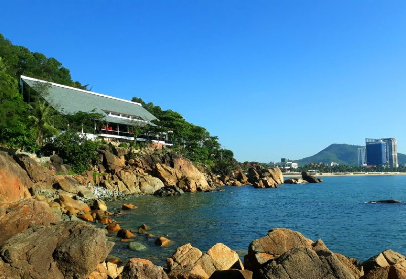 Bờ biển đ&aacute; với t&ograve;a nh&agrave; hiện đại tr&ecirc;n đồi, xa xa l&agrave; những t&ograve;a nh&agrave; cao tầng v&agrave; n&uacute;i dưới bầu trời xanh.