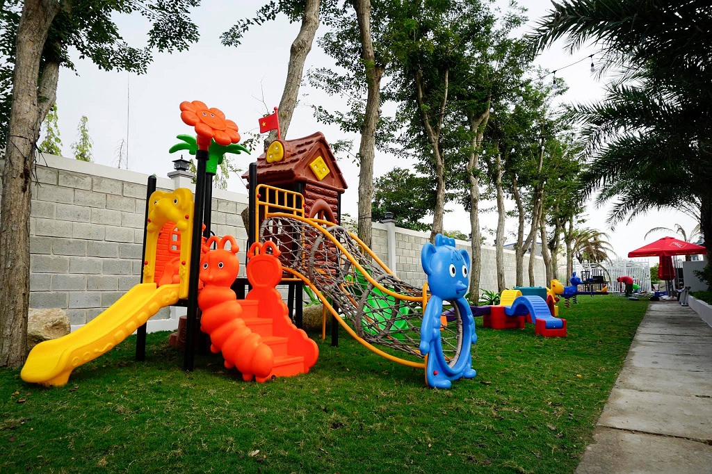 Khu vực sân chơi ngoài trời với cầu trượt, leo trèo và các thiết bị vui chơi nhiều màu sắc trên nền cỏ xanh.