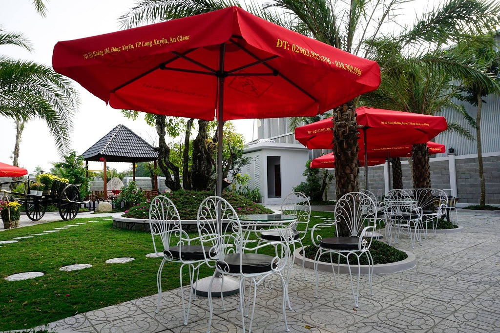 Khu vườn với bàn ghế trắng, dù đỏ, cỏ xanh và cây cọ. Có xe ngựa và chòi nghỉ nhỏ bên cạnh.