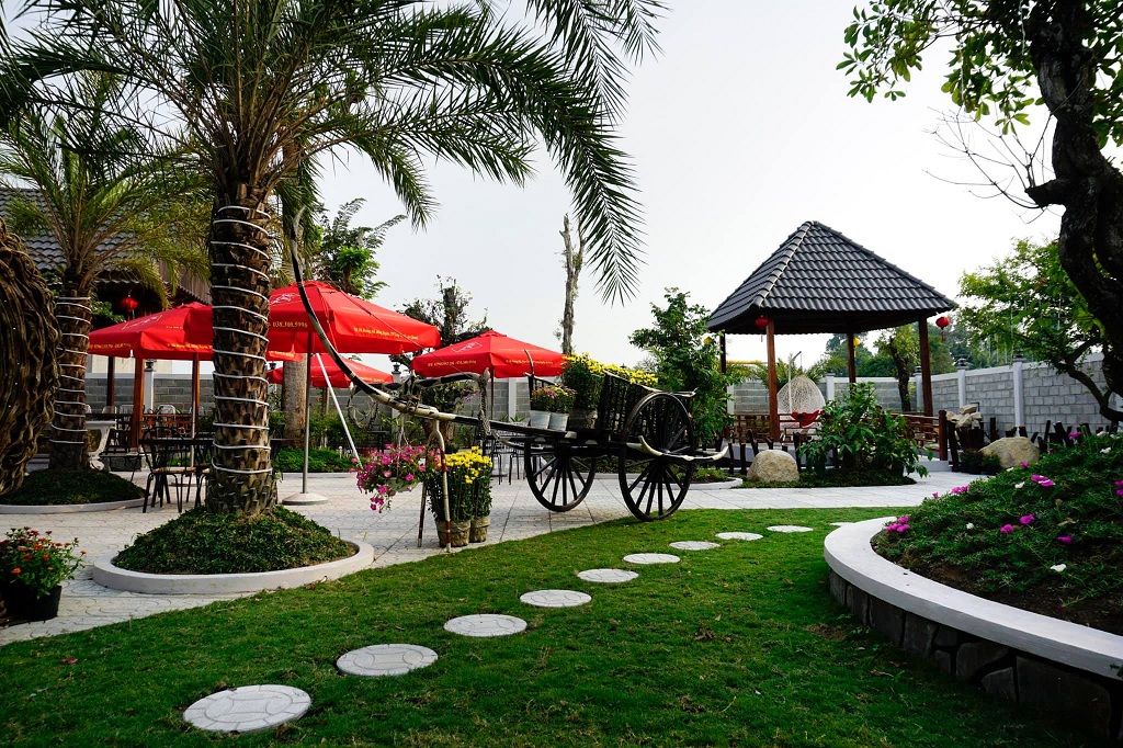 Khu vườn thoáng đãng với cây cối, lối đi, xe kéo cổ, dù đỏ và chòi nghỉ lát ngói, tạo không gian thư giãn.