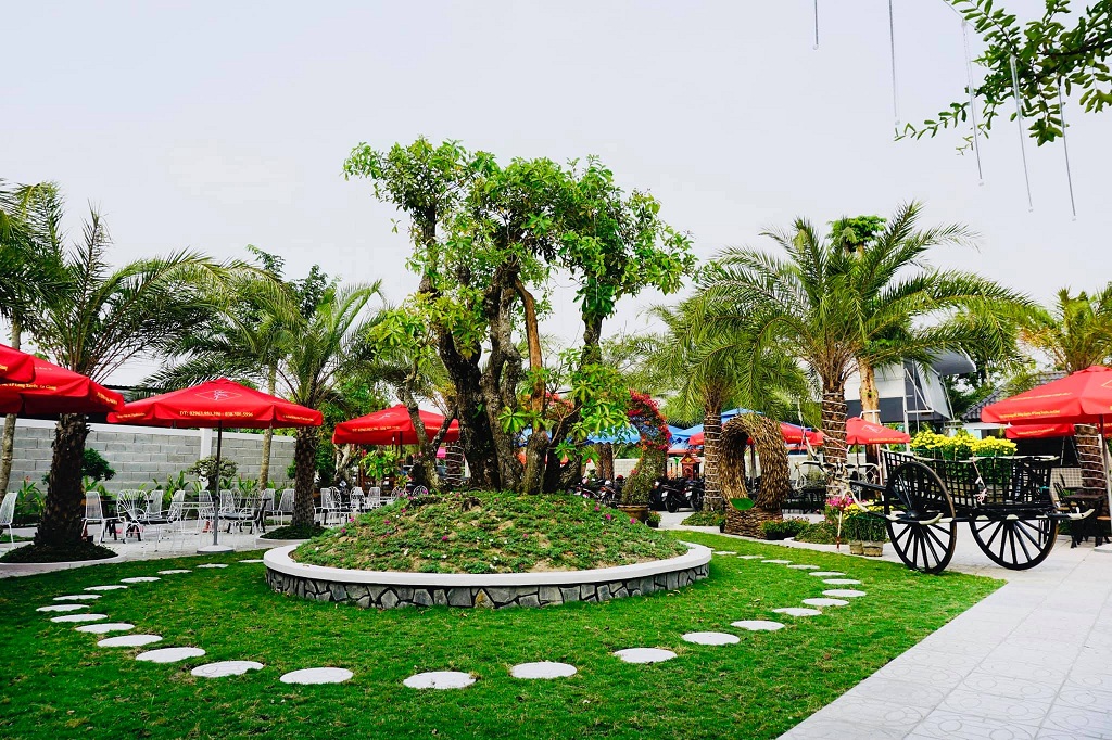 Khu vườn với cây xanh, lối đi lát đá, bàn ghế và dù đỏ, tạo không gian thư giãn ngoài trời.