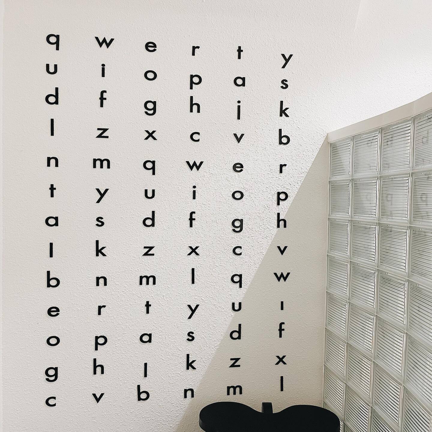 Một bức tường trắng với c&aacute;c chữ c&aacute;i đen xếp th&agrave;nh cột. C&oacute; một bức tường k&iacute;nh b&ecirc;n cạnh.