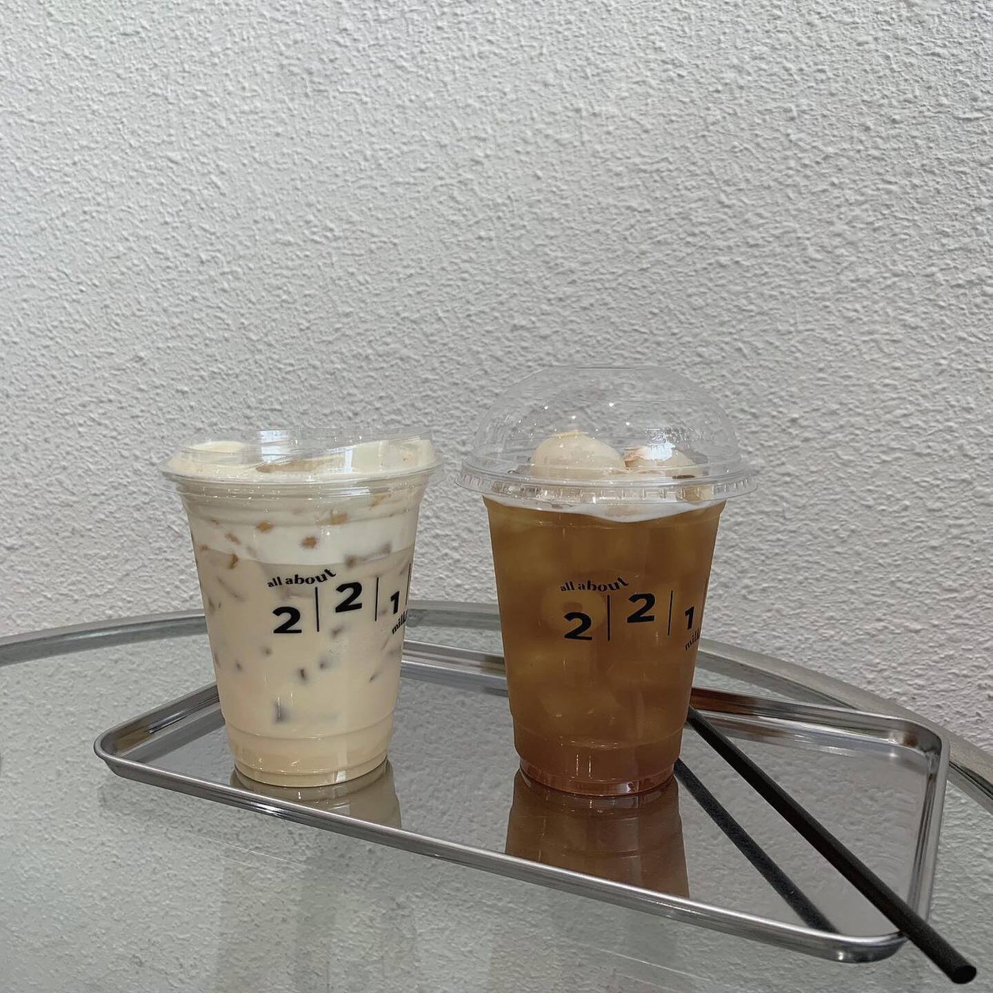 Hai ly đồ uống lạnh tr&ecirc;n khay kim loại, một ly m&agrave;u trắng sữa v&agrave; một ly m&agrave;u n&acirc;u, c&oacute; nắp nhựa trong suốt.