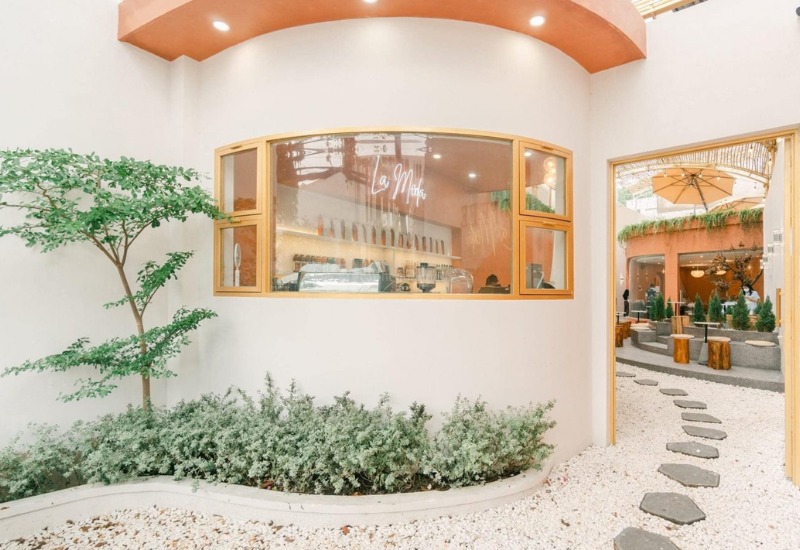 Kh&ocirc;ng gian qu&aacute;n c&agrave; ph&ecirc; với cửa sổ tr&ograve;n, c&acirc;y xanh, v&agrave; lối đi l&aacute;t đ&aacute; dẫn ra s&acirc;n vườn c&oacute; &ocirc; che nắng.