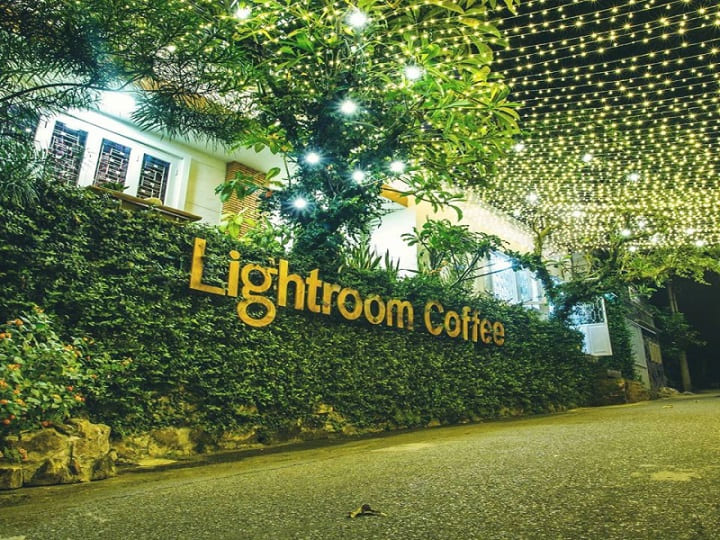 Quán cà phê với bảng hiệu "Lightroom Coffee", bên ngoài trang trí đèn và cây xanh vào ban đêm.