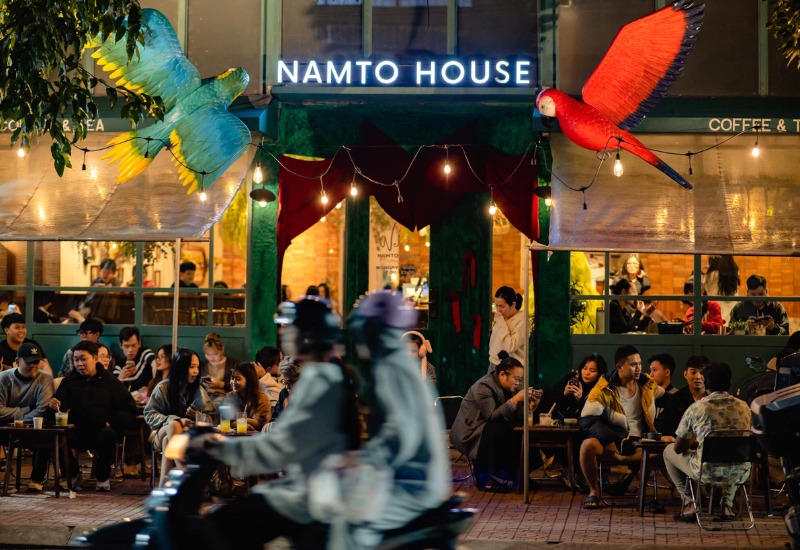 Qu&aacute;n c&agrave; ph&ecirc; đ&ocirc;ng đ&uacute;c v&agrave;o buổi tối, với hai con vẹt trang tr&iacute; lớn b&ecirc;n ngo&agrave;i v&agrave; kh&aacute;ch ngồi tr&ograve; chuyện.