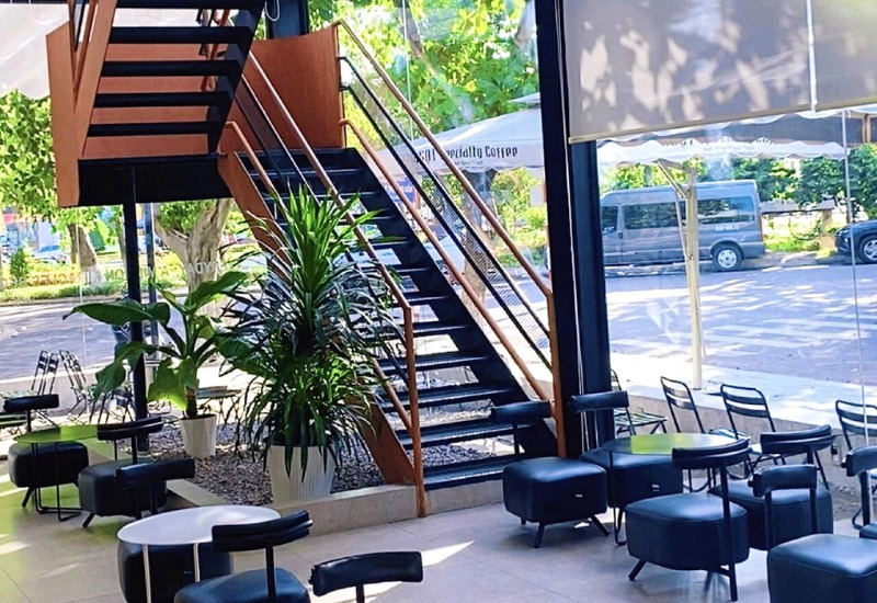 Qu&aacute;n c&agrave; ph&ecirc; với cầu thang gỗ, b&agrave;n ghế hiện đại v&agrave; c&acirc;y xanh, &aacute;nh s&aacute;ng tự nhi&ecirc;n từ cửa k&iacute;nh lớn.