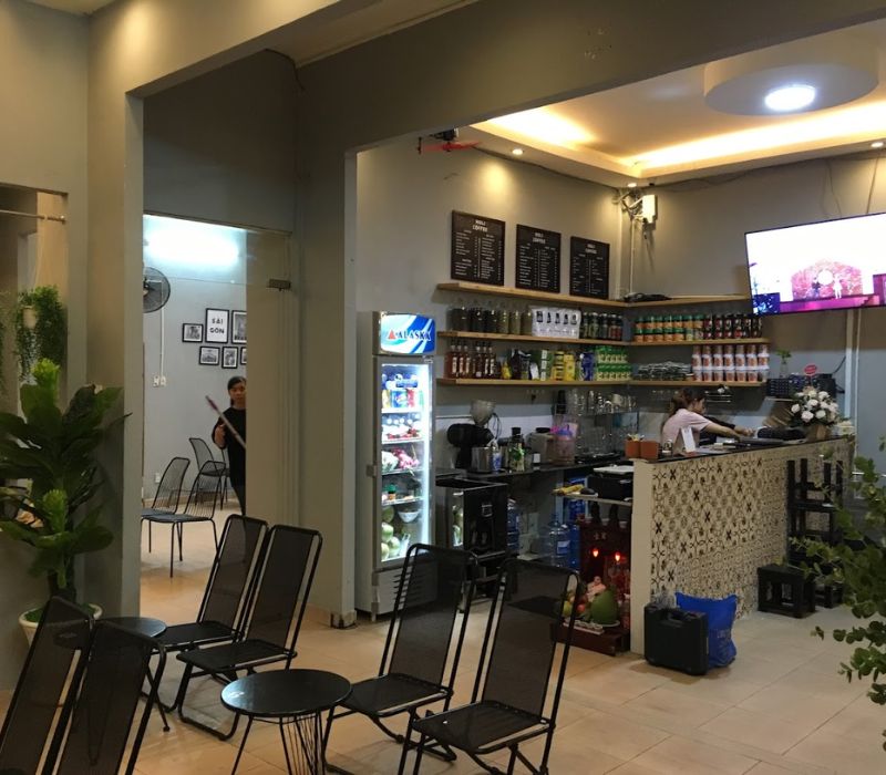 Qu&aacute;n c&agrave; ph&ecirc; với quầy pha chế, b&agrave;n ghế kim loại v&agrave; tivi treo tường, c&oacute; người đang đứng l&agrave;m việc.