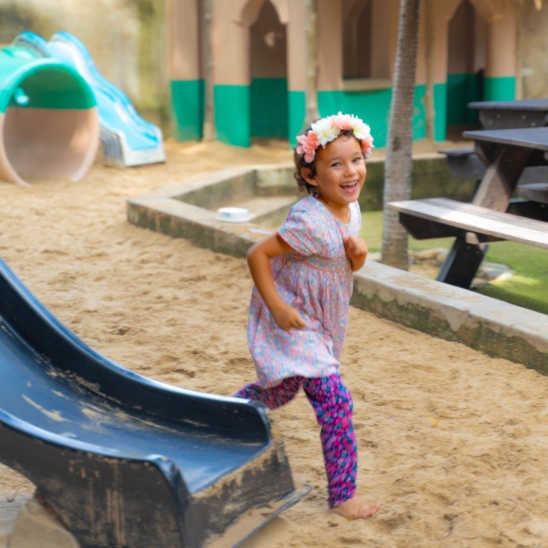 Bé gái vui chơi ở sân chơi ngoài trời với cầu trượt, đội vòng hoa trên đầu.