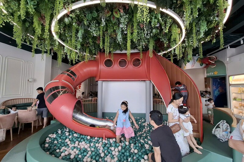 Khu vui chơi trong nh&agrave; với cầu trượt đỏ, b&oacute;ng nhựa v&agrave; c&acirc;y xanh trang tr&iacute;, c&oacute; người lớn v&agrave; trẻ em vui chơi.