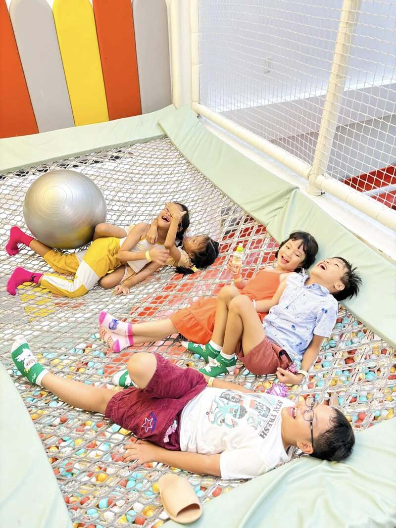 C&aacute;c em nhỏ nằm chơi vui vẻ tr&ecirc;n lưới trong khu vui chơi với quả b&oacute;ng lớn b&ecirc;n cạnh.