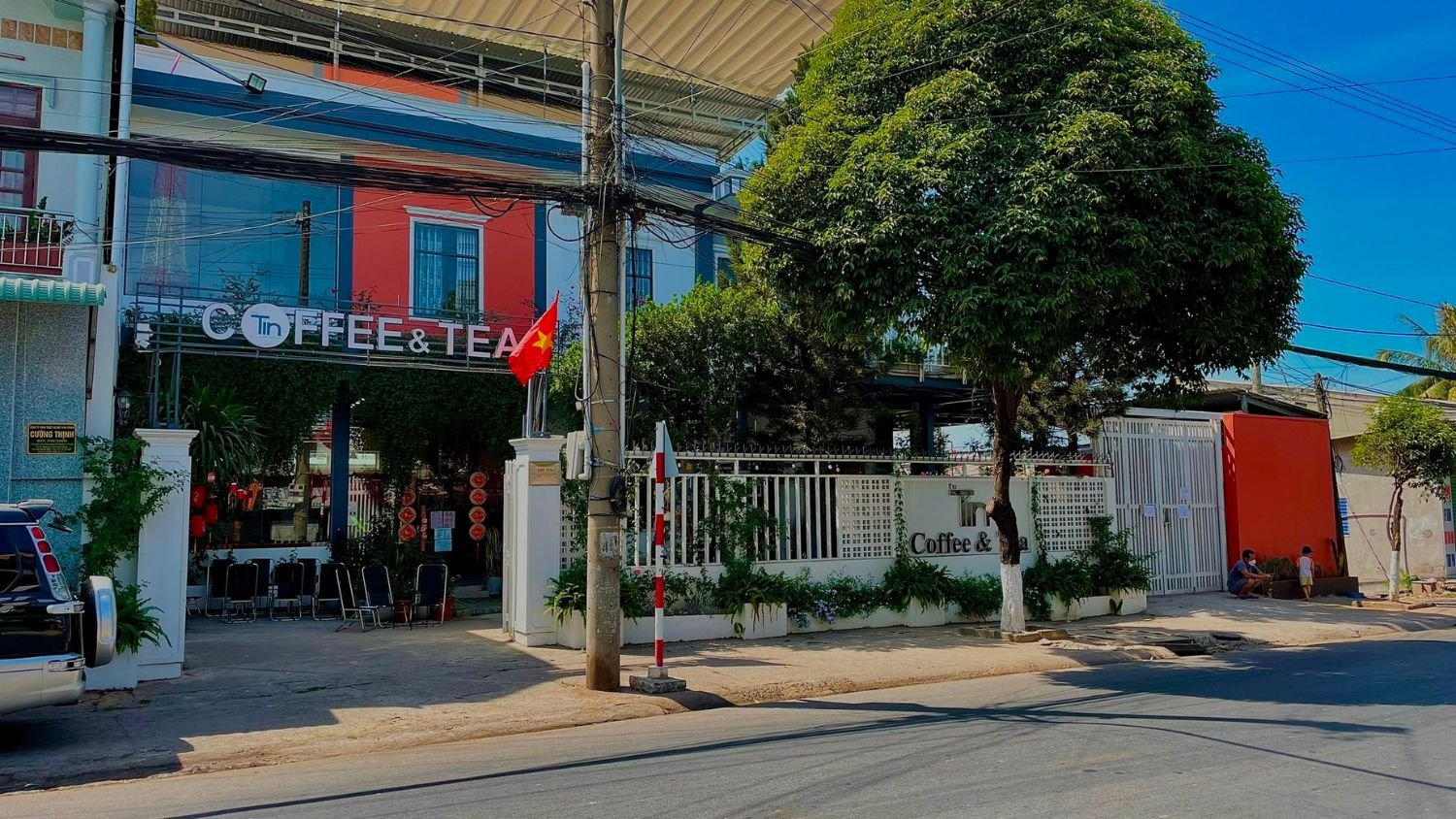 Quán cà phê và trà hiện đại, mặt tiền rộng, có cây xanh và cờ Việt Nam, trên một con đường phố yên tĩnh.