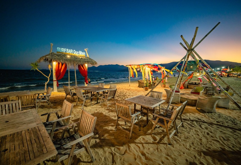 Qu&aacute;n bar b&atilde;i biển với b&agrave;n ghế gỗ, &aacute;nh s&aacute;ng lung linh, biển xanh v&agrave; n&uacute;i xa xa dưới bầu trời ho&agrave;ng h&ocirc;n.