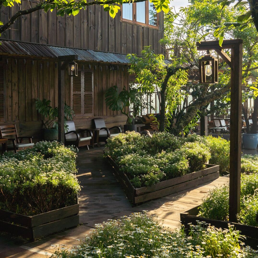 Khu vườn nhỏ với c&acirc;y xanh v&agrave; hoa trước ng&ocirc;i nh&agrave; gỗ, c&oacute; ghế ngồi thư gi&atilde;n dưới &aacute;nh nắng nhẹ.