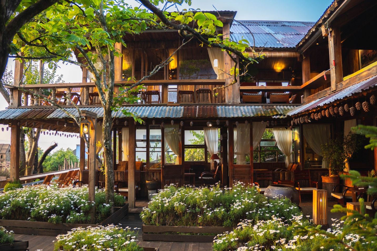 Ng&ocirc;i nh&agrave; gỗ hai tầng với hi&ecirc;n rộng, được trang tr&iacute; bằng r&egrave;m trắng v&agrave; hoa c&uacute;c trước s&acirc;n.