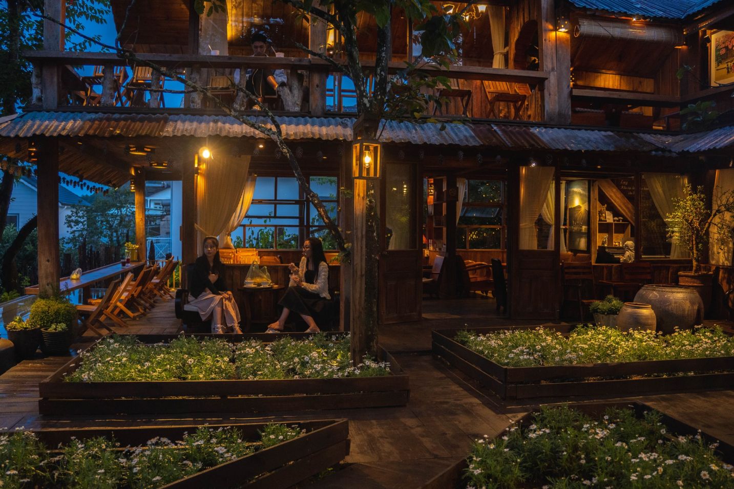 Kh&ocirc;ng gian qu&aacute;n c&agrave; ph&ecirc; bằng gỗ, &aacute;nh s&aacute;ng ấm &aacute;p, với kh&aacute;ch ngồi tr&ograve; chuyện b&ecirc;n ngo&agrave;i v&agrave;o buổi tối.