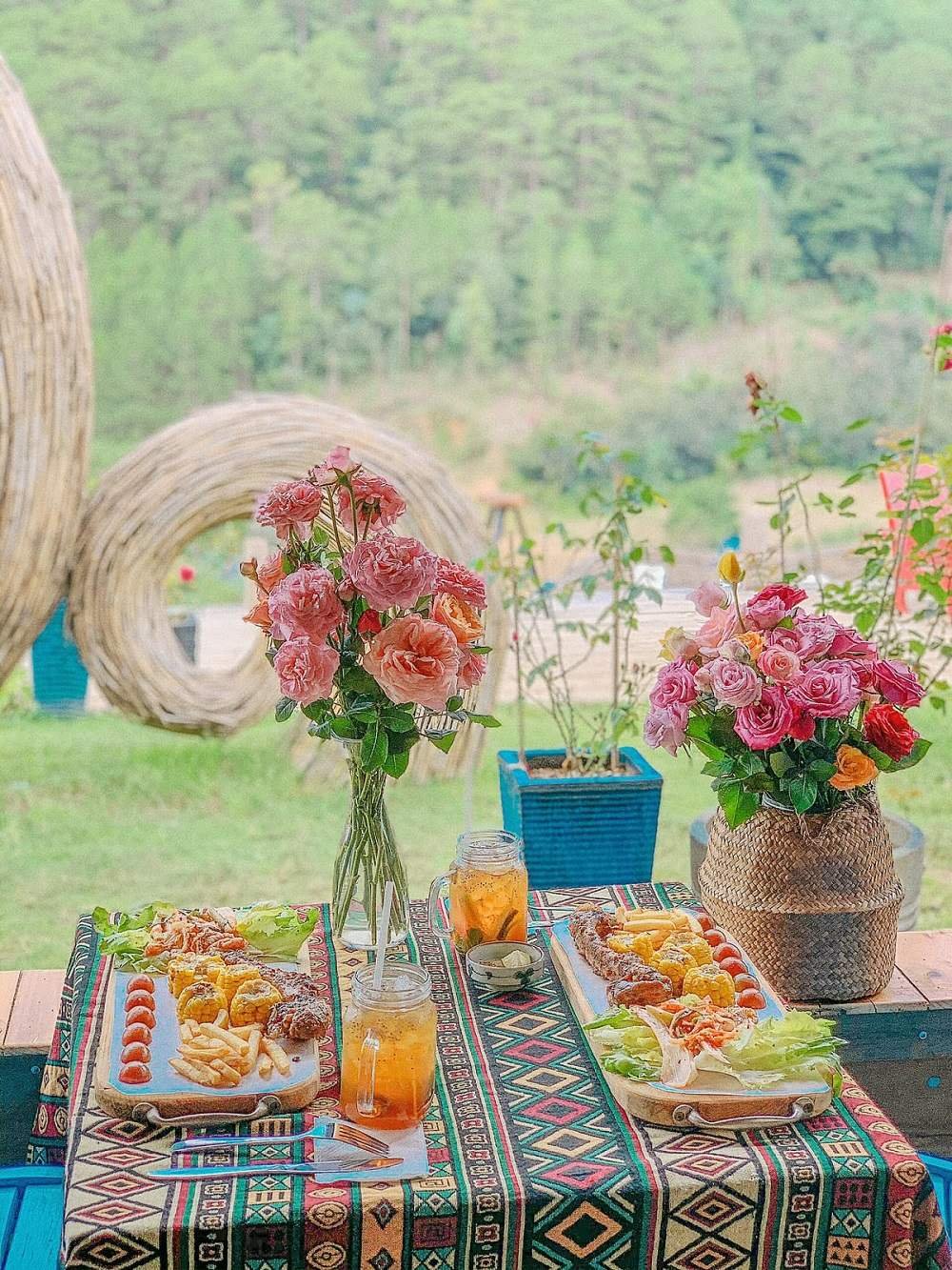 Bàn ăn ngoài trời với hoa hồng, món ăn hấp dẫn và ly trà đá trên nền thiên nhiên xanh tươi, phong cảnh thư giãn.
