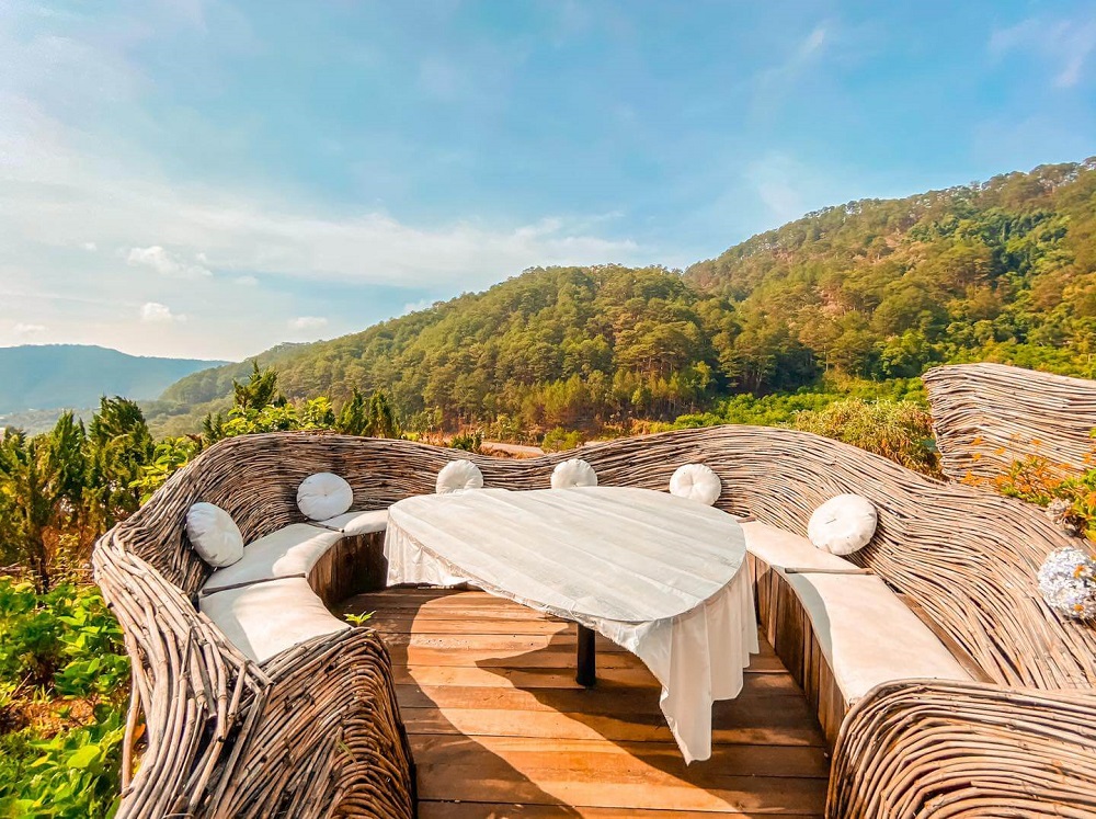 B&agrave;n ghế m&acirc;y đặt ngo&agrave;i trời, xung quanh l&agrave; cảnh n&uacute;i non xanh tươi dưới bầu trời xanh trong.