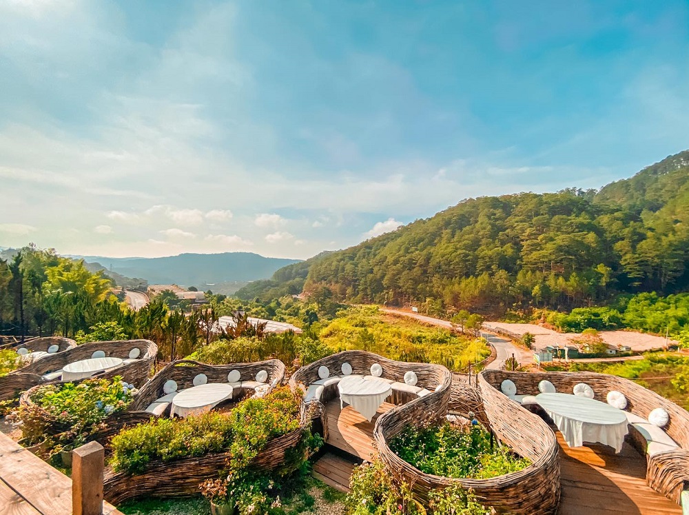 Khu vực ăn uống ngo&agrave;i trời với b&agrave;n ghế đan l&aacute;t, bao quanh l&agrave; c&acirc;y xanh v&agrave; đồi n&uacute;i dưới bầu trời xanh.