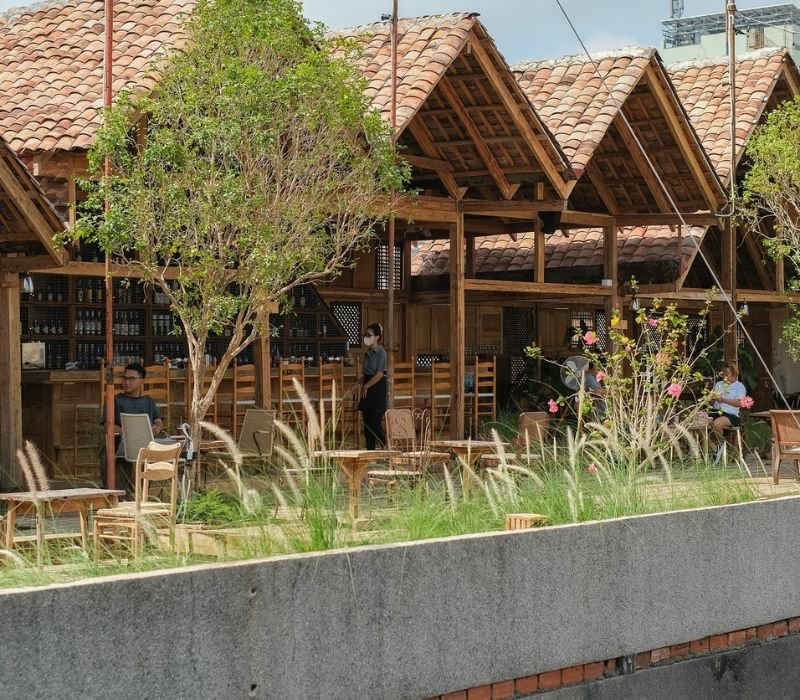 Quán cà phê sân vườn mát mẻ với kiến trúc gỗ và mái ngói, cây xanh xung quanh, khách tận hưởng không gian thư giãn.