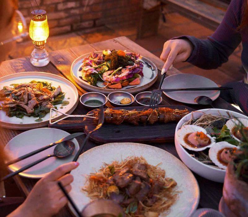 B&agrave;n ăn với nhiều m&oacute;n ăn đa dạng, c&oacute; người đang d&ugrave;ng đũa v&agrave; nĩa để thưởng thức.