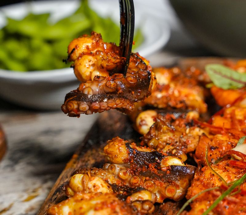 M&oacute;n bạch tuộc nướng cay tr&ecirc;n đĩa gỗ, được gắp bằng nĩa, ph&iacute;a sau l&agrave; b&aacute;t đậu xanh.