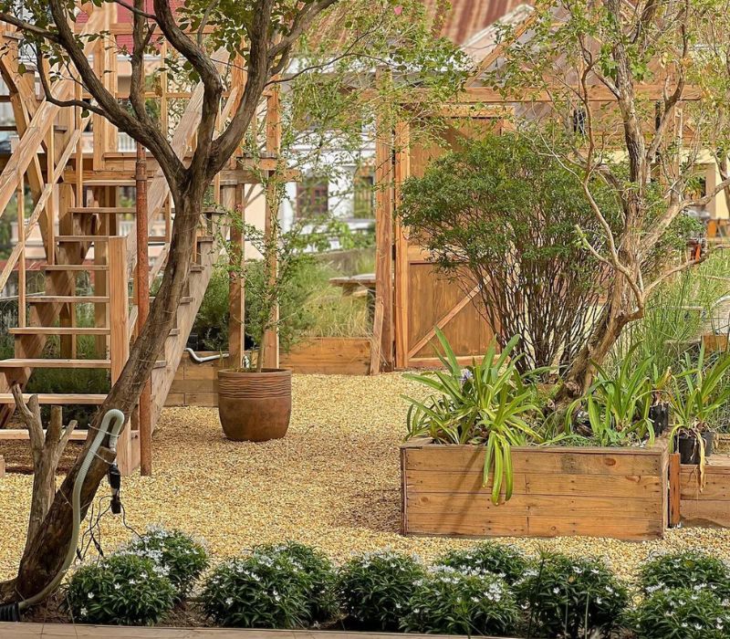 Khu vườn với c&acirc;y xanh, lối đi sỏi v&agrave; c&aacute;c chậu c&acirc;y gỗ, gần cầu thang gỗ v&agrave; nh&agrave; gỗ nhỏ.