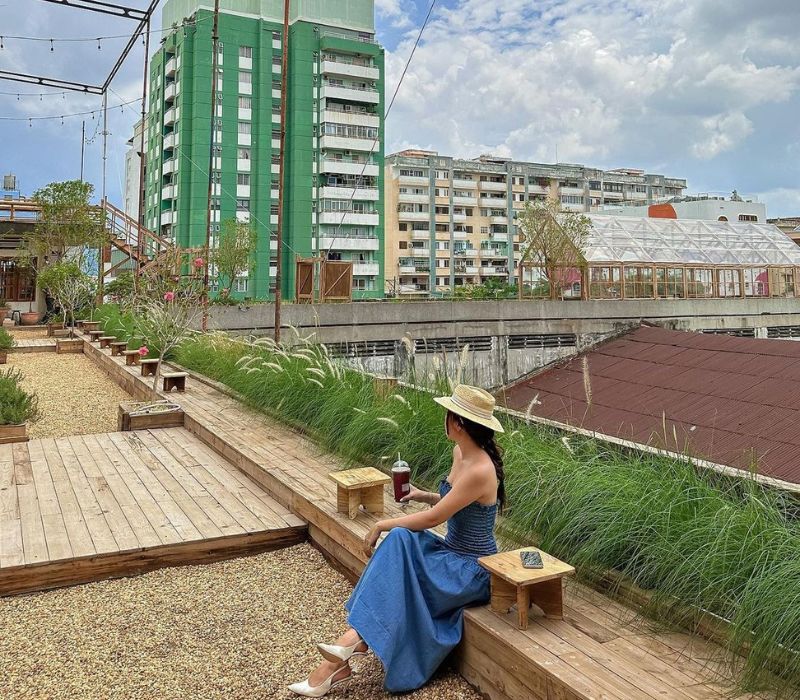 Người phụ nữ ngồi tr&ecirc;n s&acirc;n thượng c&oacute; c&acirc;y xanh, mặc v&aacute;y xanh v&agrave; đội mũ rộng v&agrave;nh, b&ecirc;n cạnh l&agrave; c&aacute;c t&ograve;a nh&agrave; cao tầng.