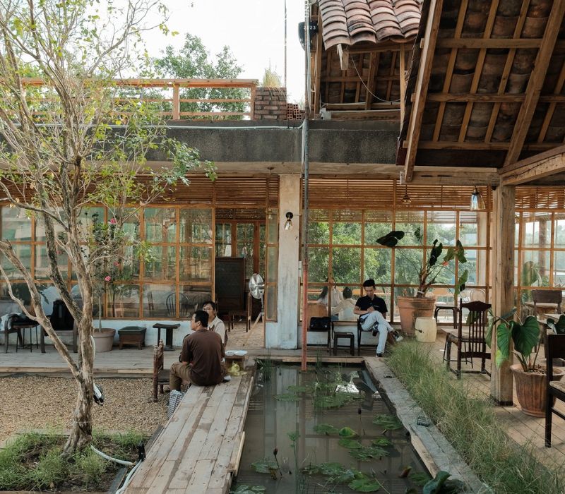 Kh&ocirc;ng gian qu&aacute;n c&agrave; ph&ecirc; ngo&agrave;i trời với m&aacute;i ng&oacute;i, hồ nước nhỏ v&agrave; c&acirc;y cối, v&agrave;i người đang ngồi thư gi&atilde;n.