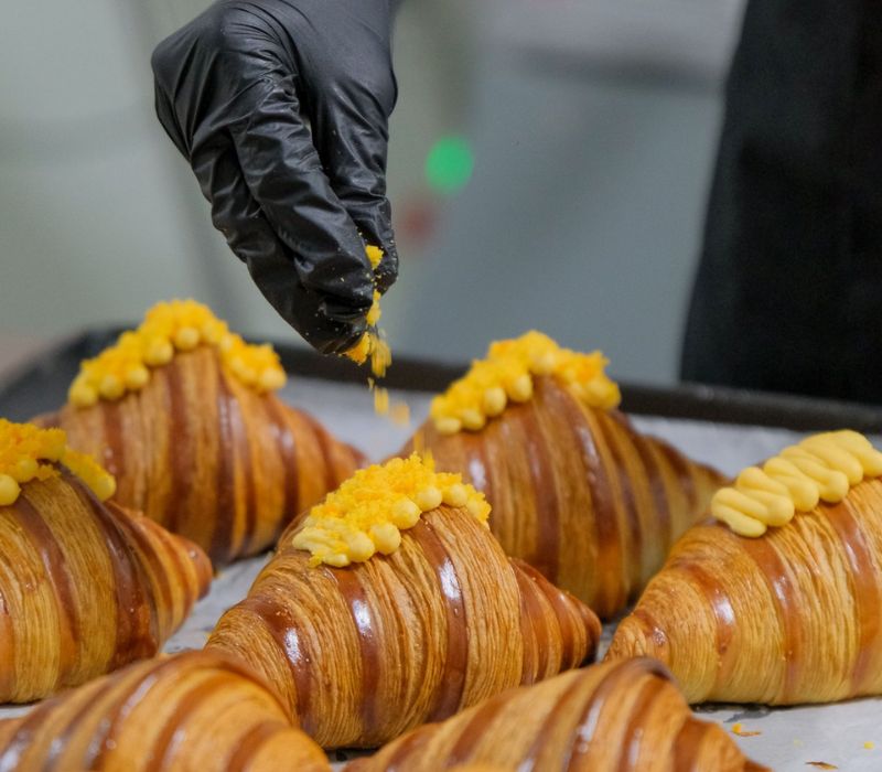 Người đeo găng tay đen trang tr&iacute; b&aacute;nh croissant với lớp phủ m&agrave;u v&agrave;ng cam tr&ecirc;n khay nướng.