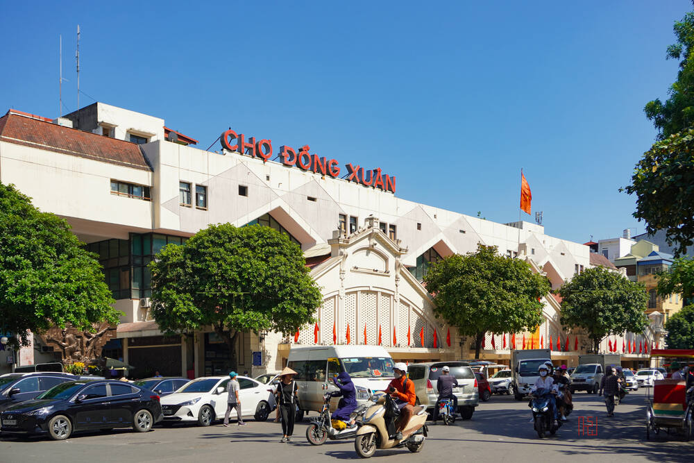 Khám Phá Phố Đường Tàu Phùng Hưng: Trải Nghiệm Cà Phê Độc Đáo Tại Hà Nội 10 Mặt tiền chợ lớn với nhiều cây xanh, xe cộ và người dân di chuyển phía trước, bầu trời trong xanh.