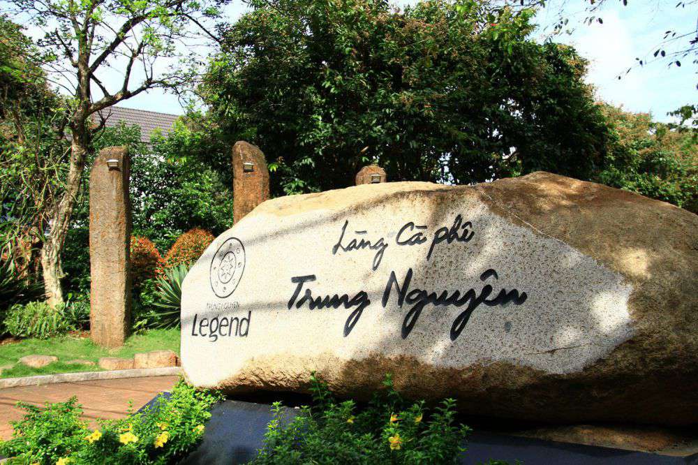 Tảng đá lớn khắc chữ "Làng Cà Phê Trung Nguyên" trong khuôn viên xanh mướt với cây cối xung quanh.