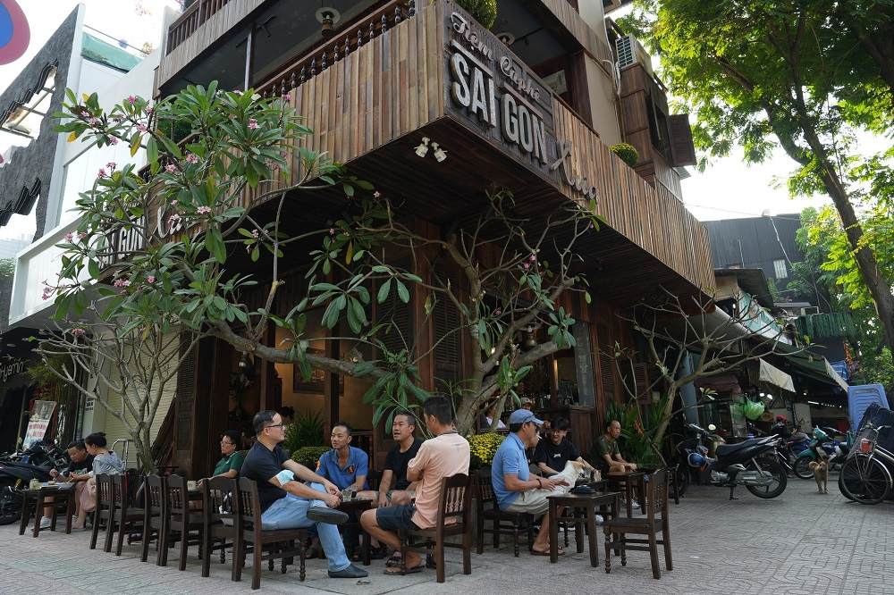 12 Quán Cafe Sài Gòn Xưa Hấp Dẫn Nhất định Phải Thử 3 Quán cà phê góc phố với nhiều cây xanh, khách ngồi uống nước, trò chuyện dưới tán cây.