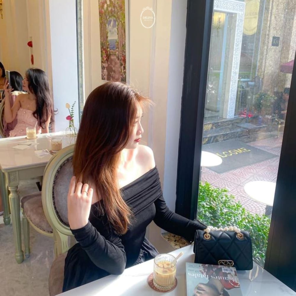 Khám Phá 10+ Quán Cafe Đẹp Nhất Long Biên: Thỏa Sức “Sống Ảo”, Thưởng Thức Đồ Uống Ngon 14 Người phụ nữ ngồi bên cửa sổ quán cà phê, tay cầm túi xách, nhìn ra ngoài, ánh nắng chiếu vào.