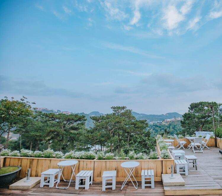 Qu&aacute;n c&agrave; ph&ecirc; ngo&agrave;i trời với b&agrave;n ghế trắng, c&acirc;y xanh v&agrave; bầu trời xanh ở Đ&agrave; Lạt.