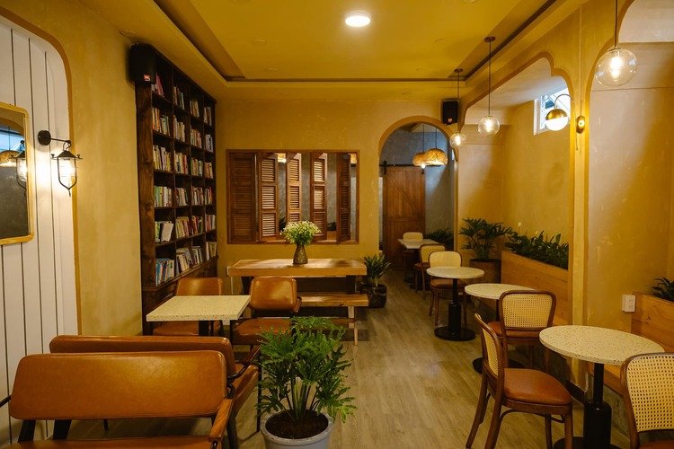 Qu&aacute;n c&agrave; ph&ecirc; ấm c&uacute;ng với b&agrave;n ghế gỗ, kệ s&aacute;ch v&agrave; c&acirc;y xanh trang tr&iacute;.