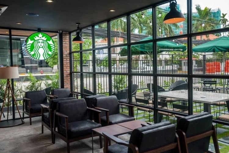 Qu&aacute;n c&agrave; ph&ecirc; với nội thất hiện đại, c&oacute; cửa sổ lớn nh&igrave;n ra ngo&agrave;i, b&agrave;n ghế gỗ v&agrave; biểu tượng thương hiệu nổi bật.