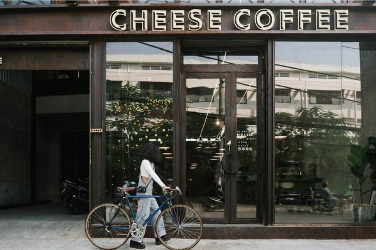 Người đẩy xe đạp đứng trước cửa qu&aacute;n c&agrave; ph&ecirc; c&oacute; t&ecirc;n "Cheese Coffee", mặt tiền k&iacute;nh trong suốt.
