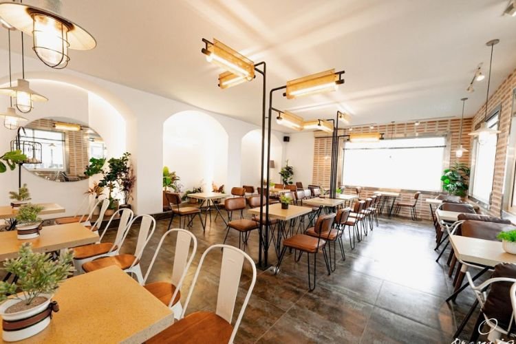 Qu&aacute;n c&agrave; ph&ecirc; với b&agrave;n ghế gỗ, &aacute;nh s&aacute;ng ấm &aacute;p, c&acirc;y xanh trang tr&iacute; v&agrave; cửa sổ lớn.