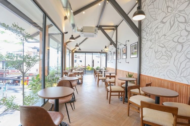 Qu&aacute;n c&agrave; ph&ecirc; s&aacute;ng sủa với b&agrave;n ghế gỗ, tường k&iacute;nh lớn v&agrave; đ&egrave;n treo, trang tr&iacute; c&acirc;y xanh v&agrave; tranh ảnh.