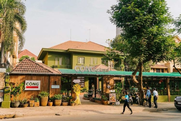 Qu&aacute;n c&agrave; ph&ecirc; với kiến tr&uacute;c xanh m&aacute;t, c&oacute; bảng hiệu v&agrave; nhiều c&acirc;y cảnh trước cửa, người đi bộ tr&ecirc;n vỉa h&egrave;.