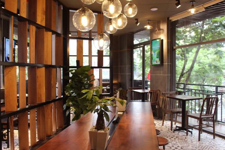 Qu&aacute;n c&agrave; ph&ecirc; với b&agrave;n gỗ d&agrave;i, c&acirc;y trang tr&iacute; v&agrave; đ&egrave;n trần, cửa sổ k&iacute;nh lớn nh&igrave;n ra cảnh c&acirc;y xanh b&ecirc;n ngo&agrave;i.