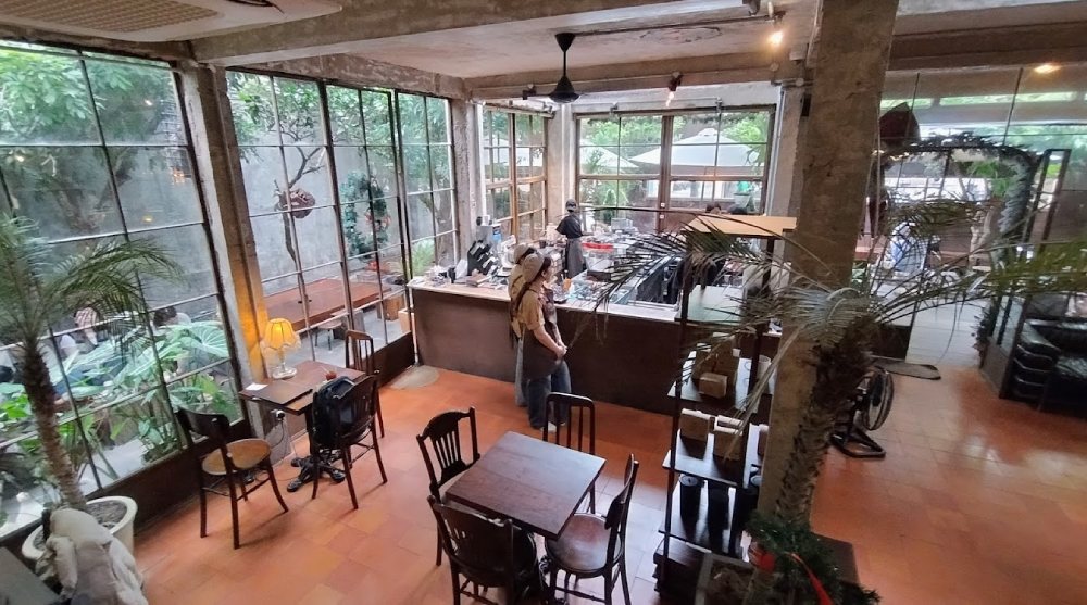 Top 11+ Quán Cafe Đẹp Tại Đà Nẵng Không Thể Bỏ Qua Năm 2025 18 Quán cà phê với thiết kế mở, có bàn ghế gỗ và cây xanh trang trí, một người đang đứng gần quầy pha chế.