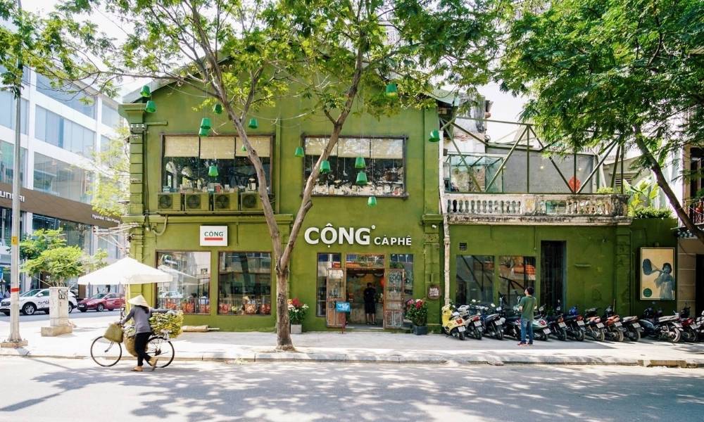Khám Phá 15 Quán Cafe Yên Tĩnh ở Quận 1: Không Gian Thoải Mái, Thức Uống Tuyệt Hảo 11 Quán cà phê màu xanh lá cây bên đường, phía trước có xe đạp và xe máy, cây xanh che bóng mát.