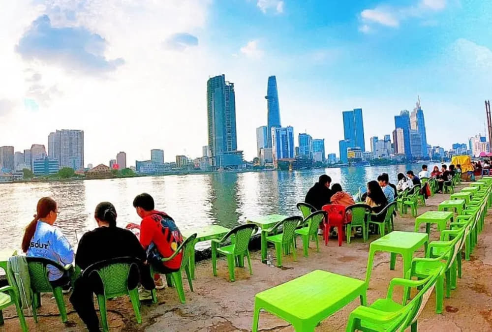 Khám Phá Cafe Bờ Kè Nóc Hầm Thủ Thiêm: Địa Điểm Thư Giãn và Ngắm Cảnh Tuyệt Vời 12 Người ngồi thưởng thức cảnh hoàng hôn bên sông với tòa nhà cao tầng ở phía xa, bàn ghế xanh xếp dọc bờ.