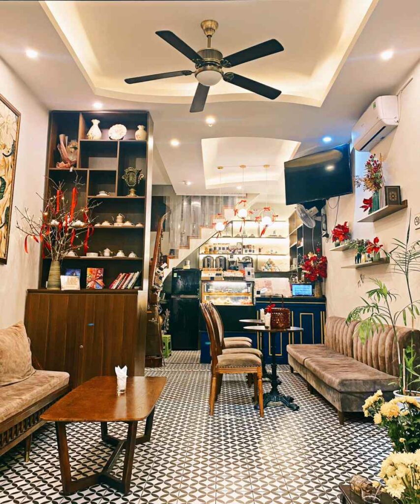 Khám Phá 10+ Quán Cafe Đẹp Nhất Long Biên: Thỏa Sức “Sống Ảo”, Thưởng Thức Đồ Uống Ngon 9 Quán cà phê với nội thất ấm cúng, bàn ghế gỗ, quạt trần, kệ sách và trang trí cây cảnh.