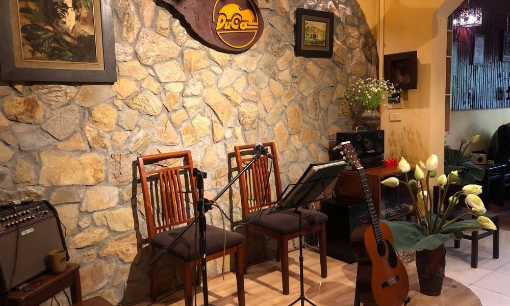 Khám Phá Top 6 Quán Cà Phê Trịnh Hấp Dẫn Tại Cầu Giấy 14 Hai ghế gỗ, micro, và đàn guitar trước bức tường đá, trang trí bằng tranh và bình hoa trong quán cà phê.