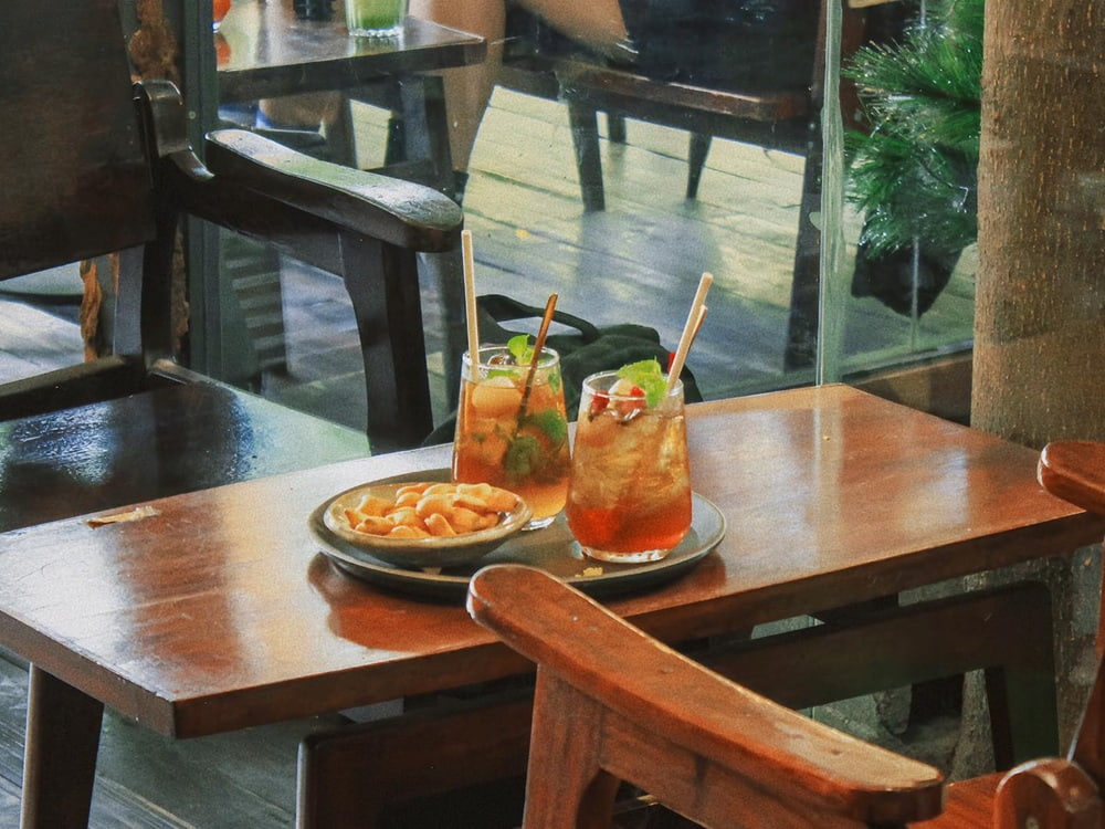 Khám Phá Cú Trên Cây Coffee: Không Gian Đà Lạt Giữa Lòng Sài Gòn 8 Hai ly trà đá và đĩa đồ ăn nhẹ trên bàn gỗ cạnh cửa sổ, trong không gian quán cà phê ấm cúng.